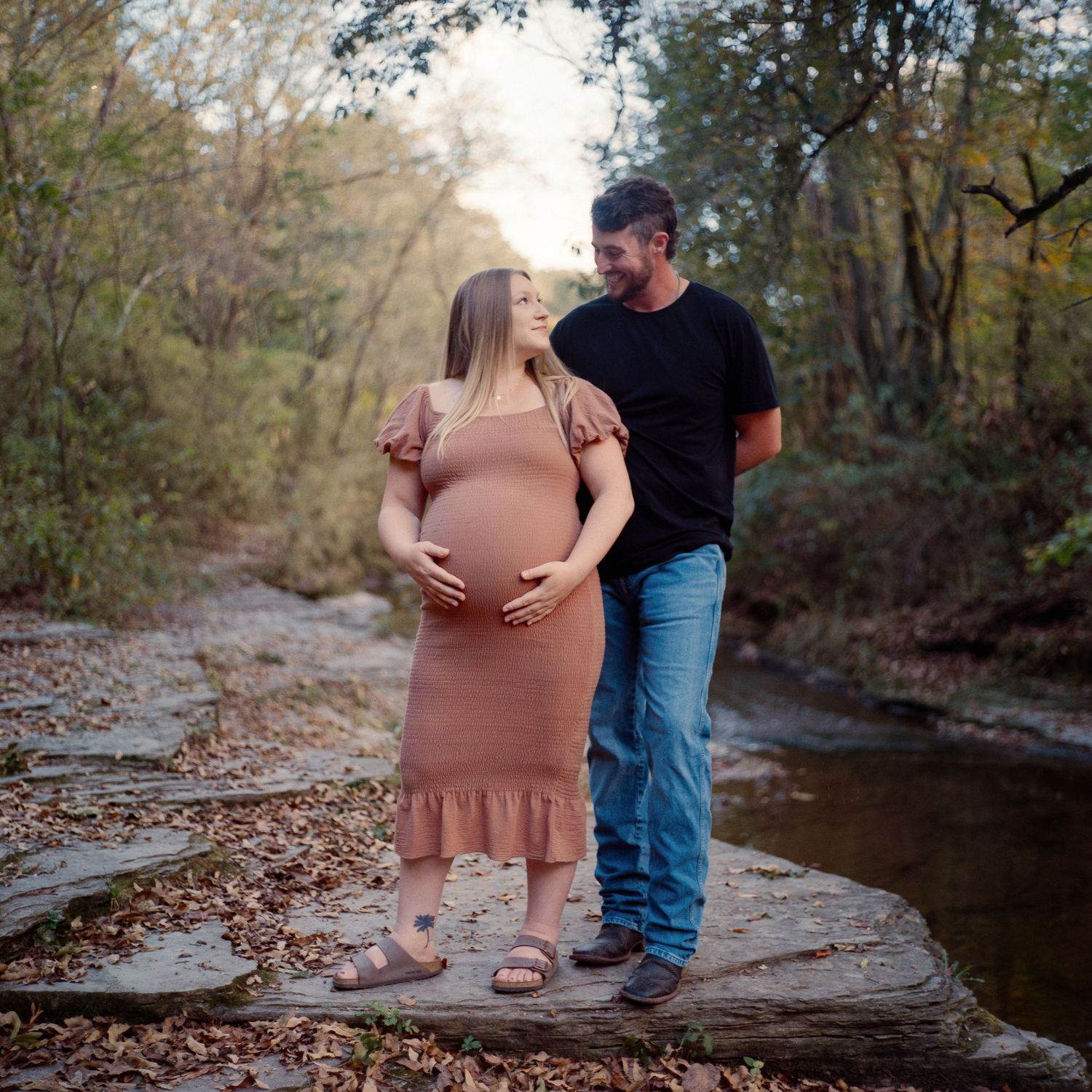Couple sharing a tender moment outdoors on rocks by a creek, woman in pink maternity dress cradling baby bump.