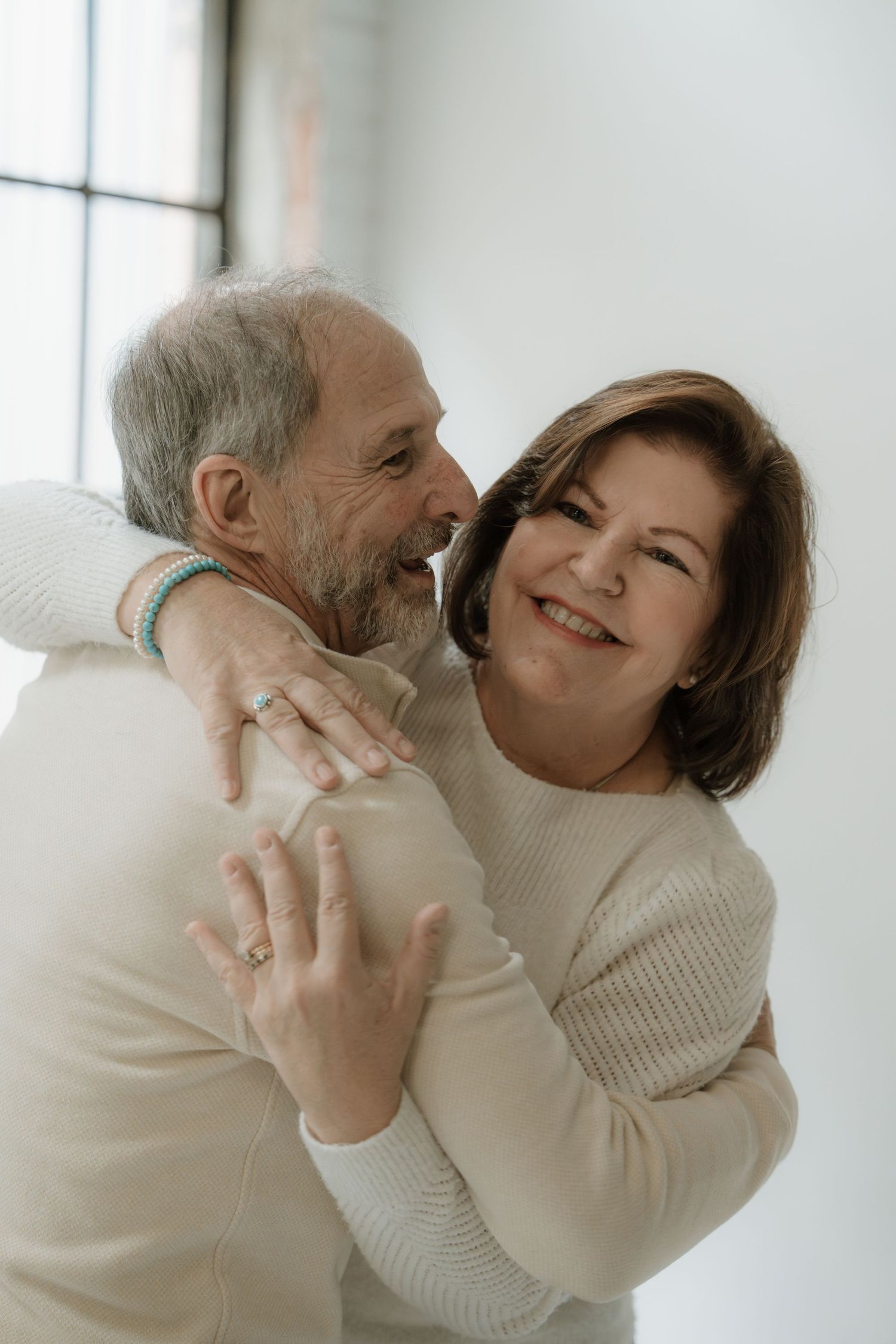 Happy older couple embracing and smiling, wearing matching cream sweaters, sharing a tender romantic moment together.