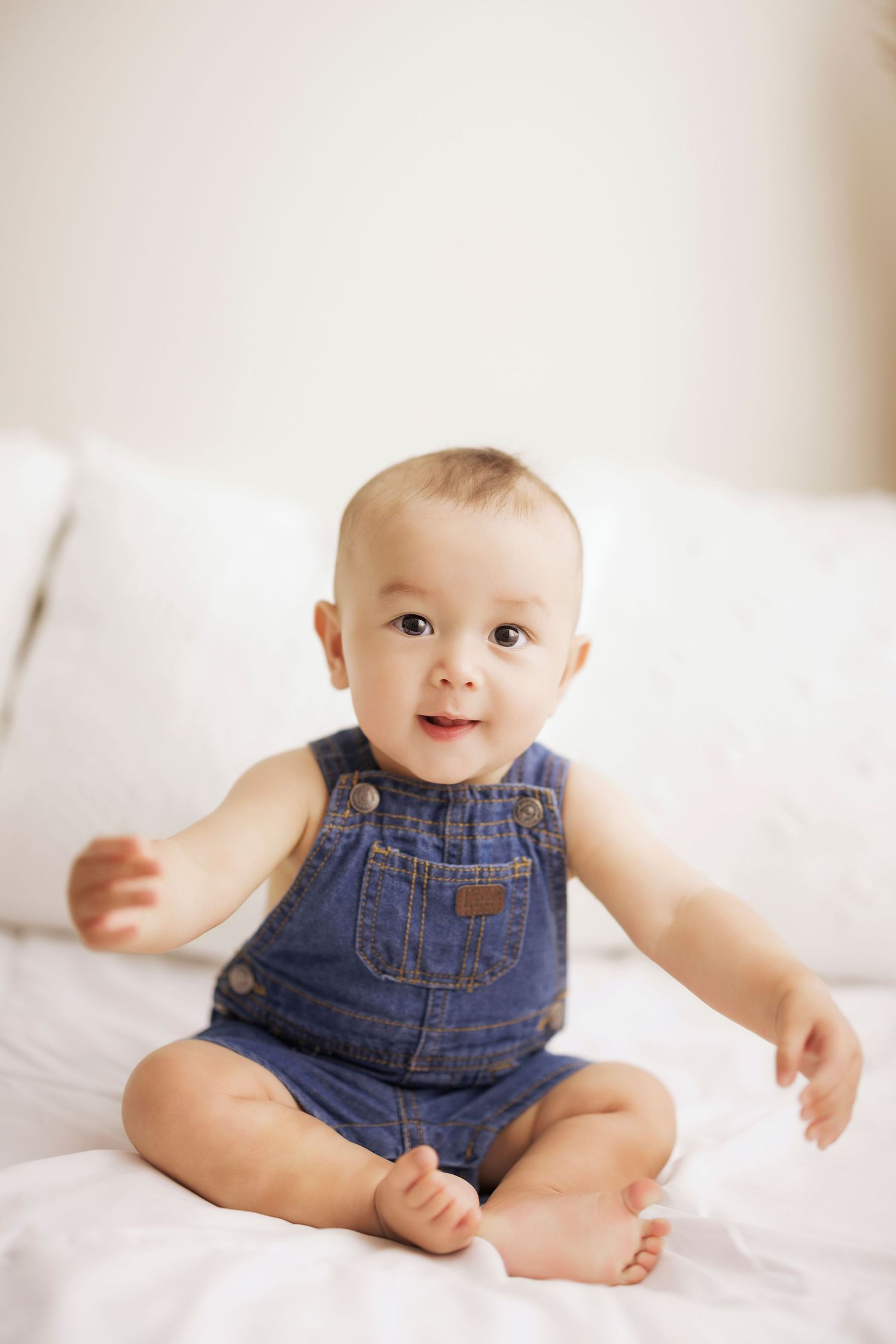 Adorable baby in denim overalls sitting on white bed, smiling with arms outstretched.