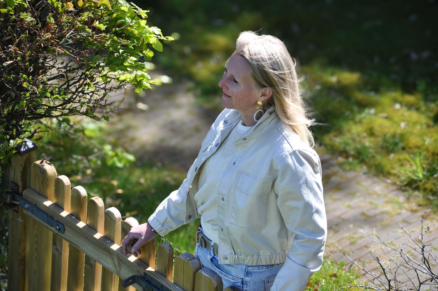 Blonde woman in light jacket standing by wooden fence in sunny garden.