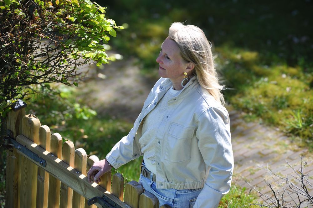 Blonde woman in light jacket standing by wooden fence in sunny garden.