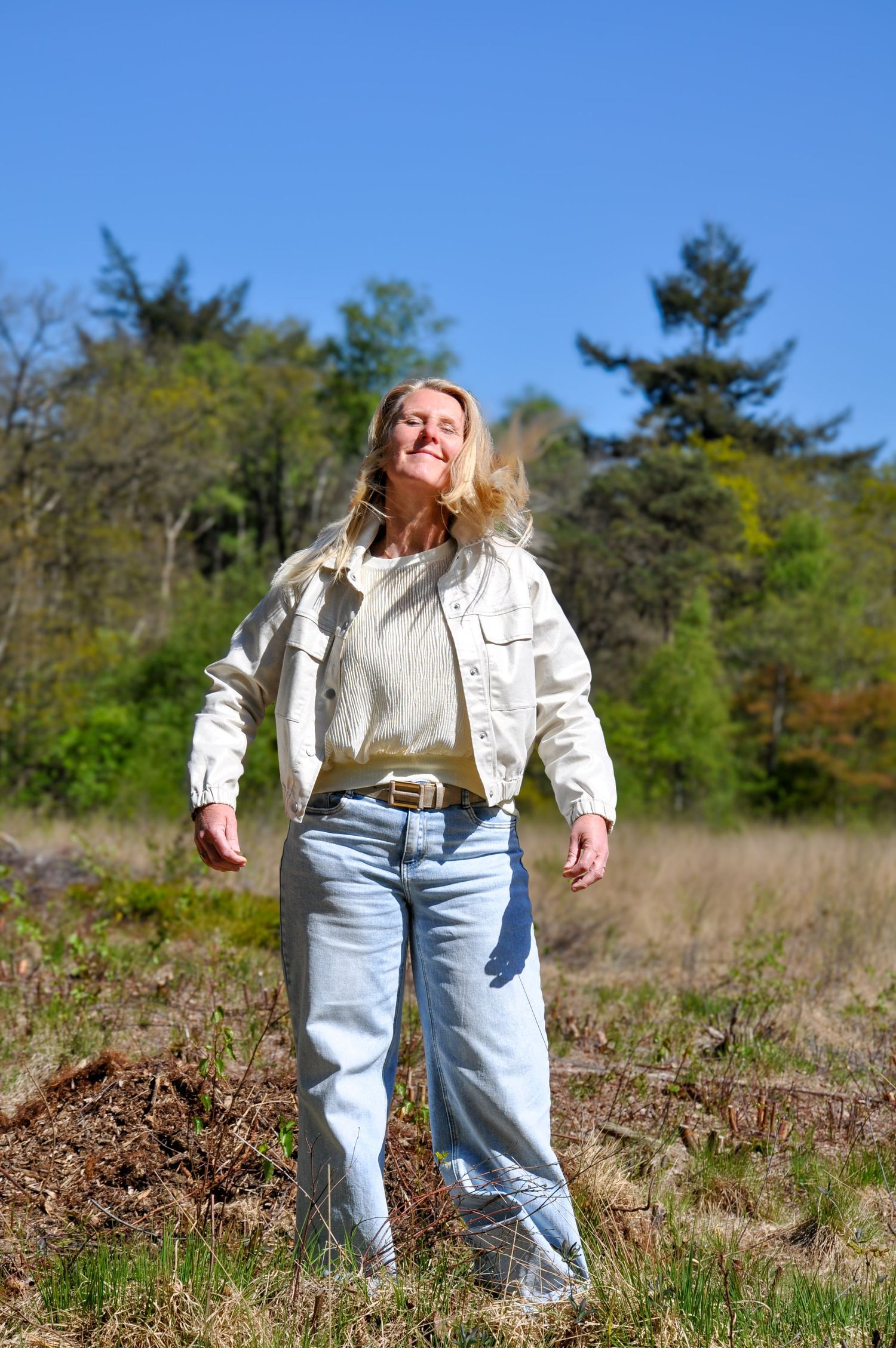 Blonde woman in white jacket and jeans enjoying sunshine in a natural meadow landscape.