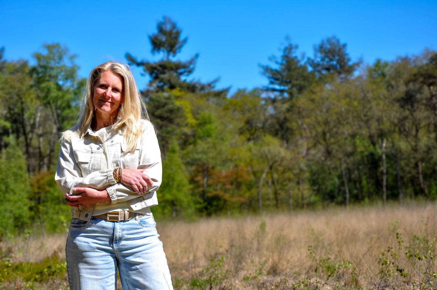 Smiling blonde woman in white jacket standing in a sunny open meadow with trees.