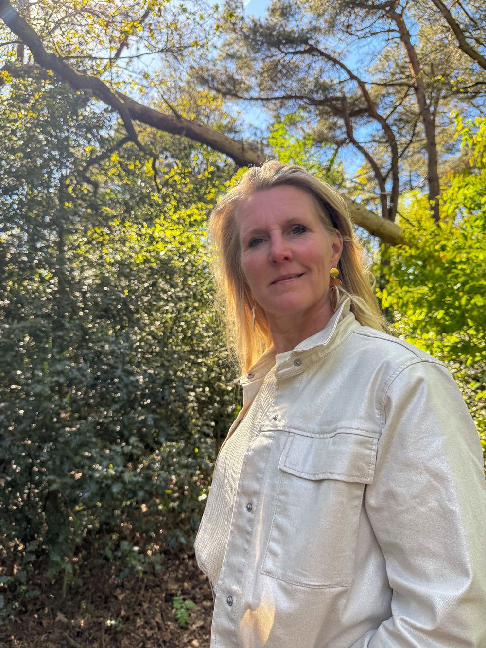Blonde woman in white denim jacket standing among lush green shrubs under blue sky.
