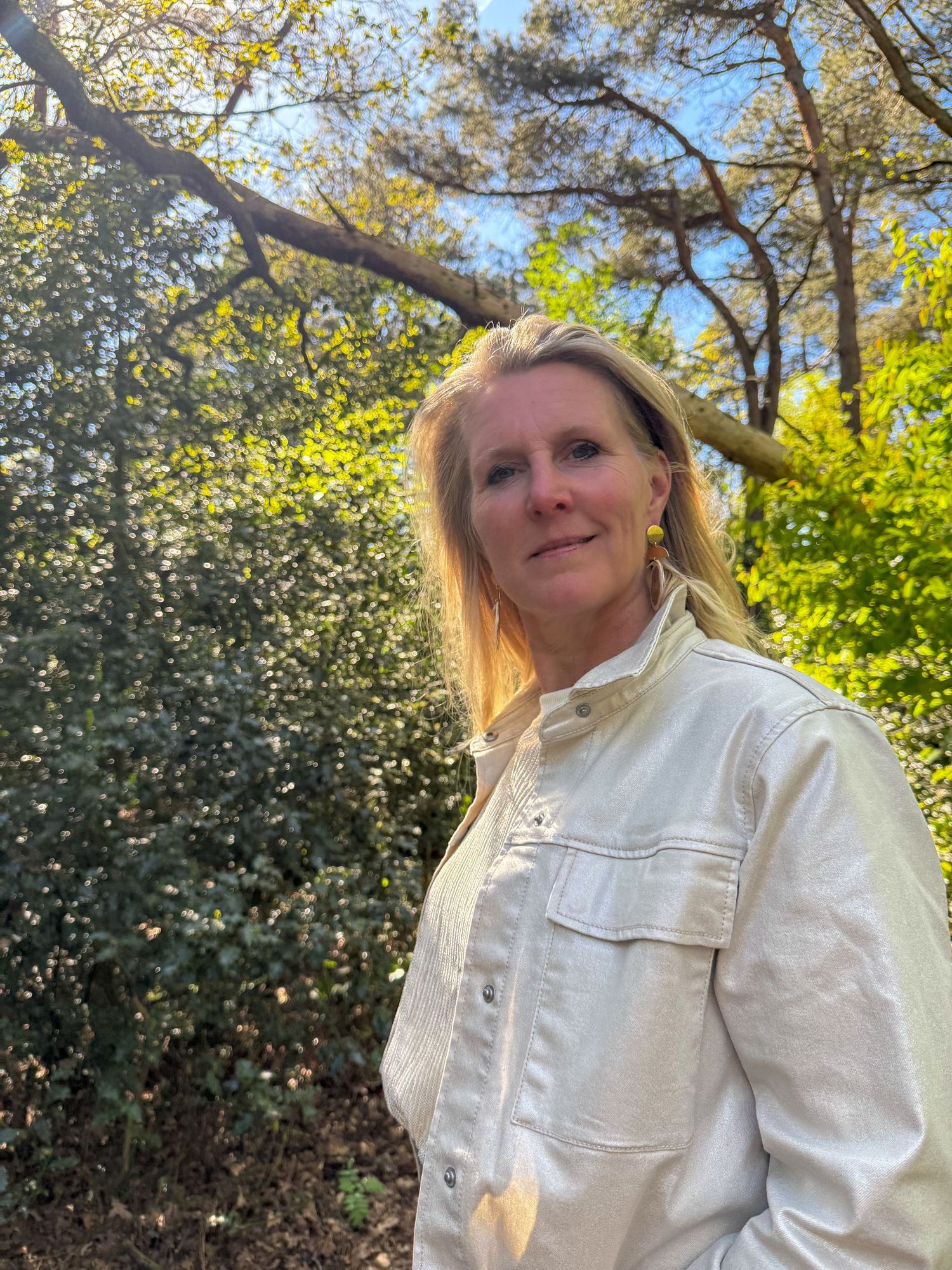 Blonde woman in white denim jacket standing among lush green shrubs under blue sky.
