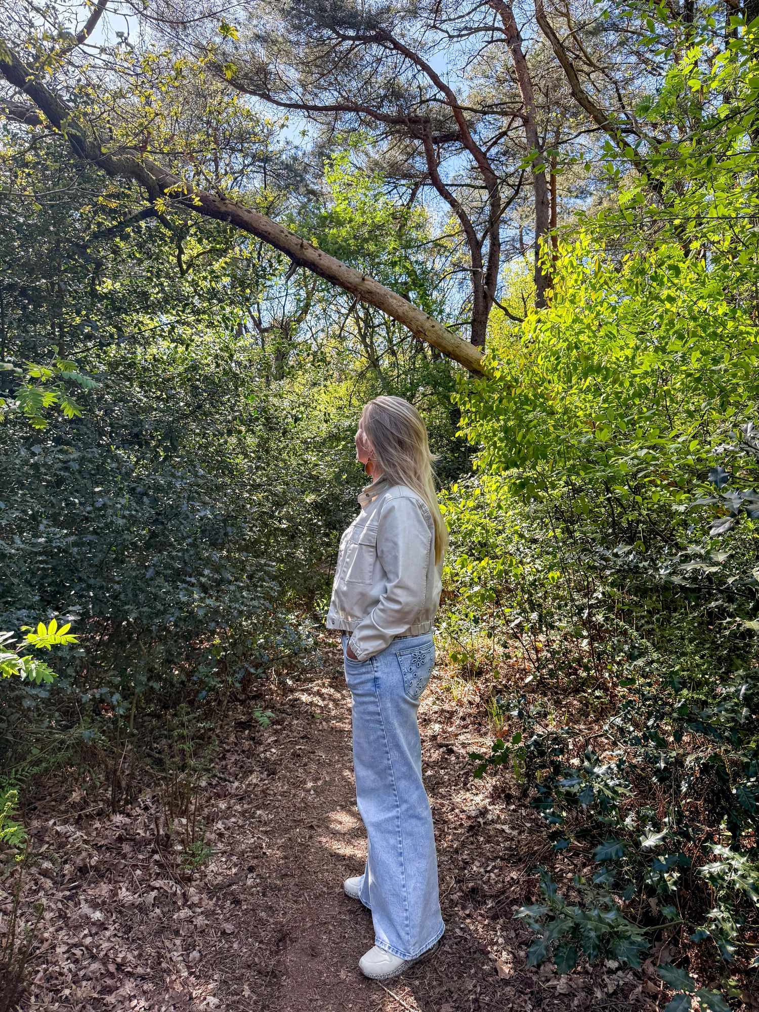 Woman with blonde hair in light jeans and jacket standing on forest trail surrounded by lush green trees.