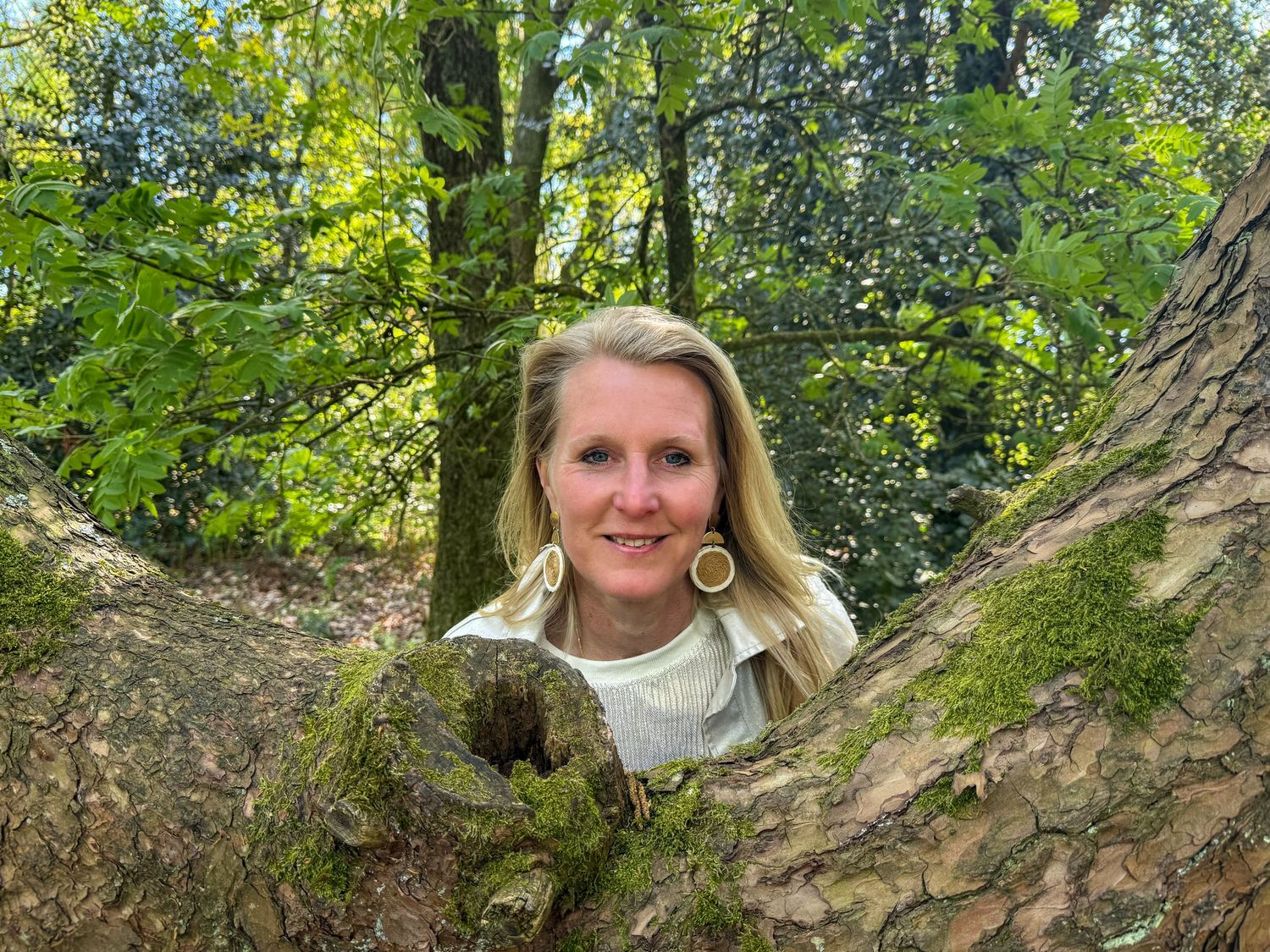Blonde woman peering through mossy tree branches in a sunlit green forest setting.