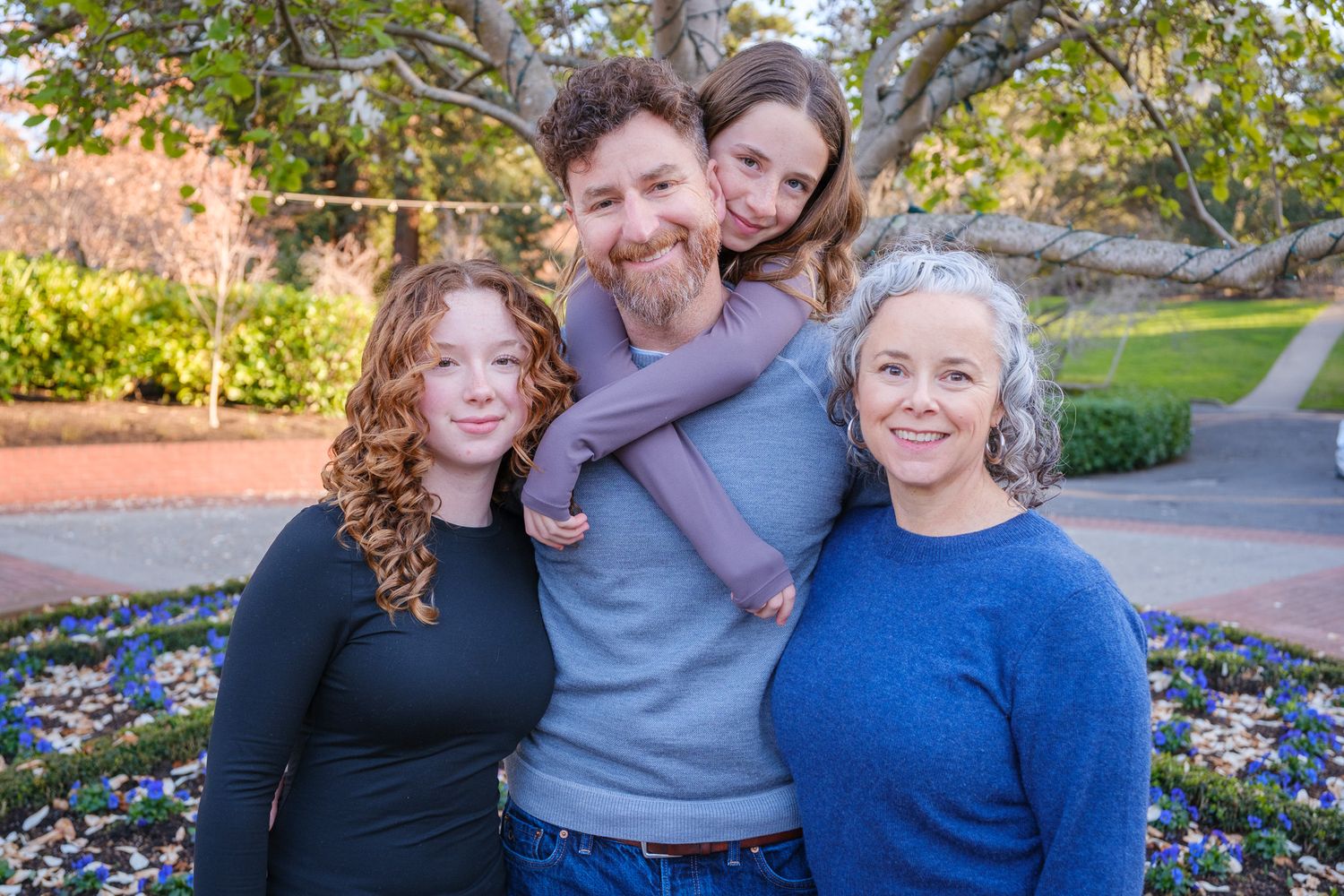Family of four smiling together outdoors in a garden, with colorful flowers and green trees in the background.