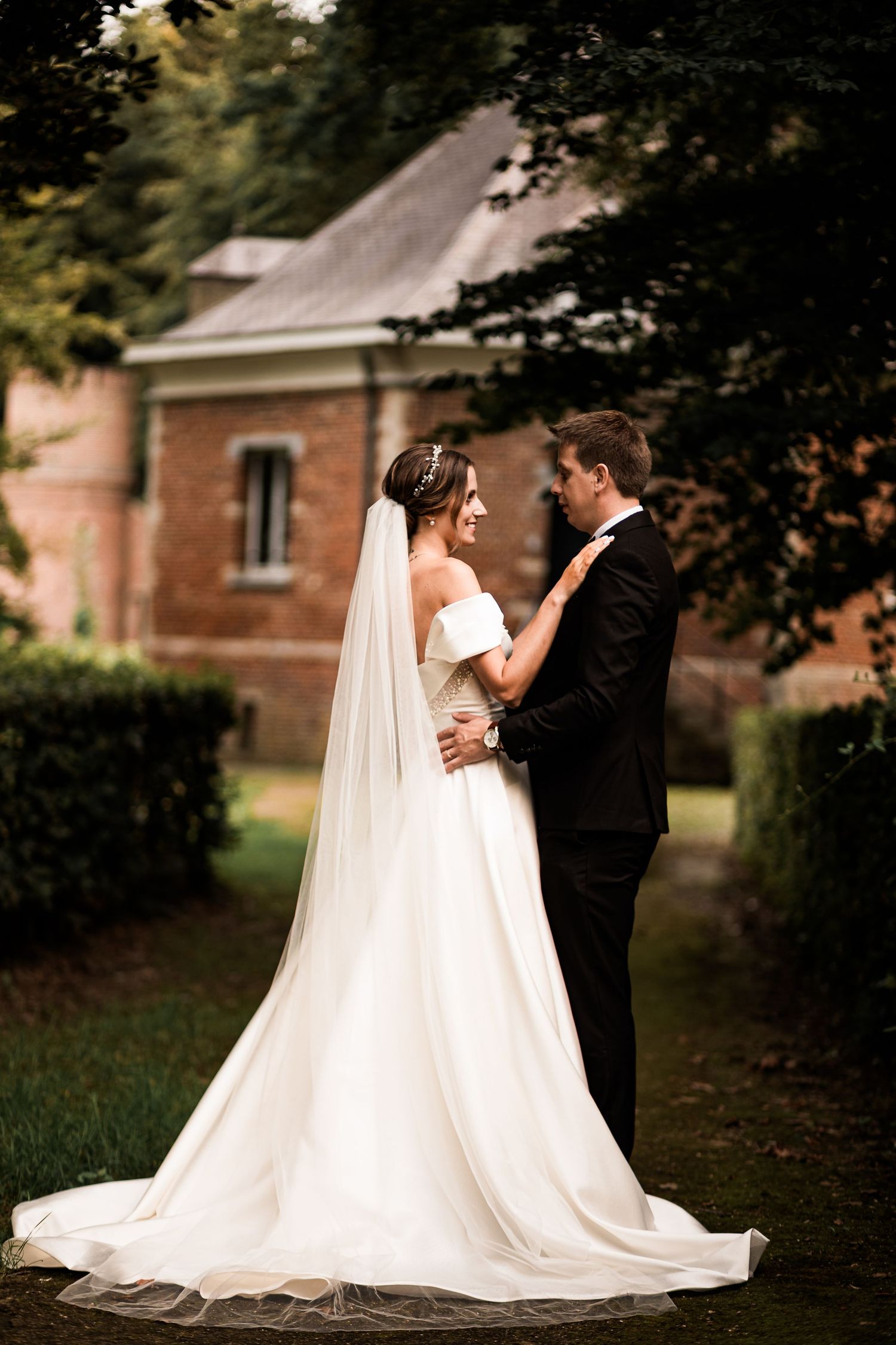 Bride in white gown and veil embraces groom in black tuxedo outdoors near brick building surrounded by lush greenery.