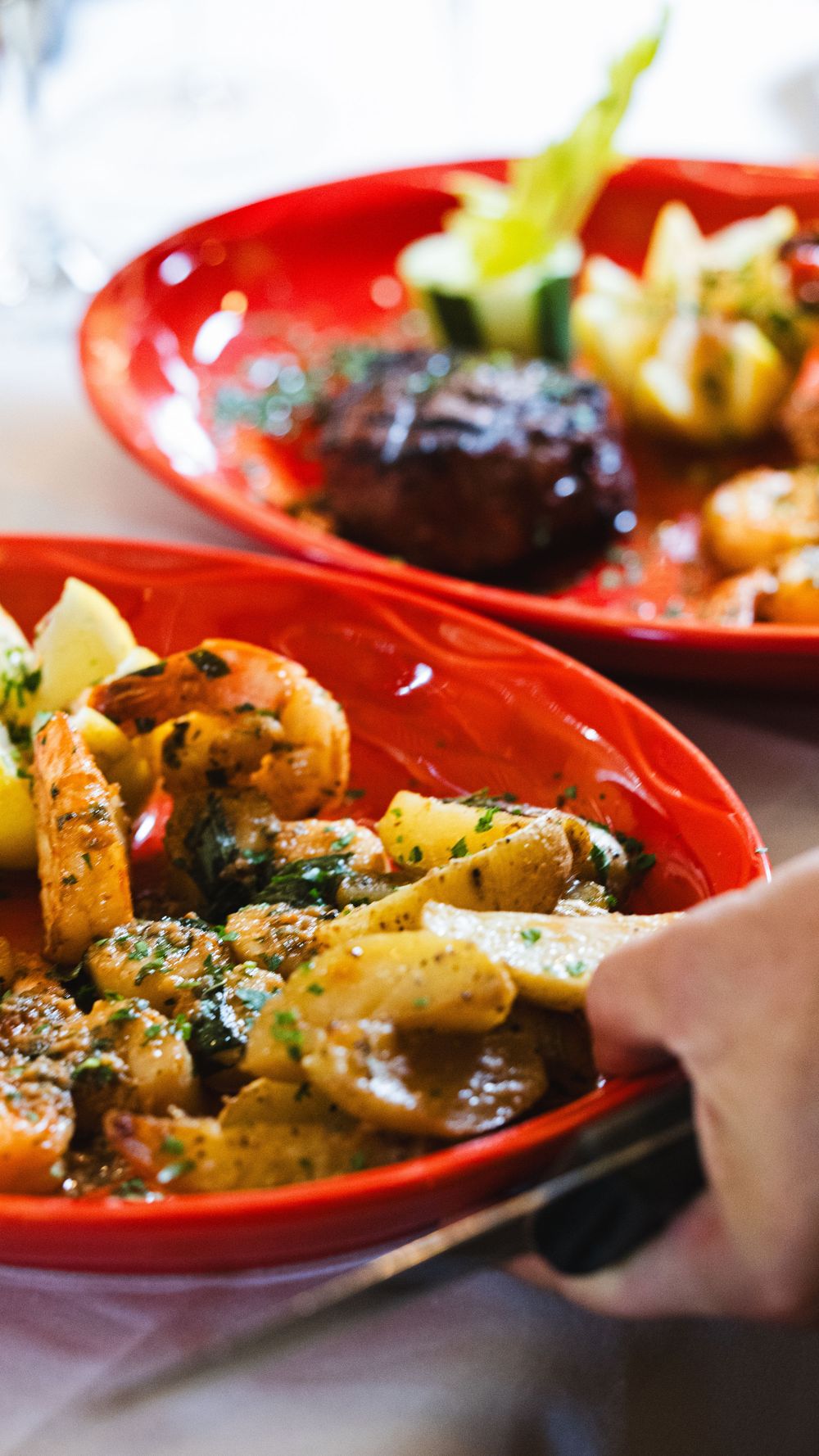 Sauteed shrimp with herbs and potatoes served in a red bowl alongside a filet mignon dish.