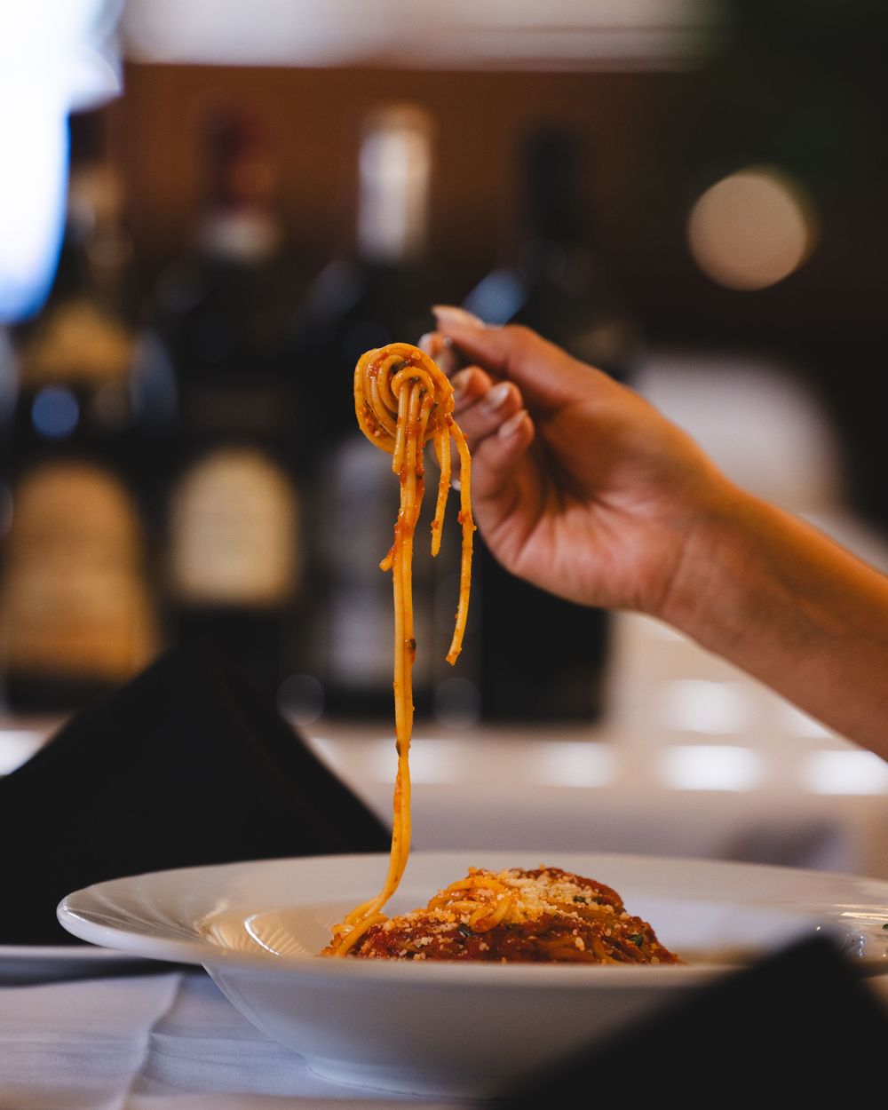 Hand lifting a forkful of spaghetti with meat sauce from a white bowl in a dimly lit restaurant.