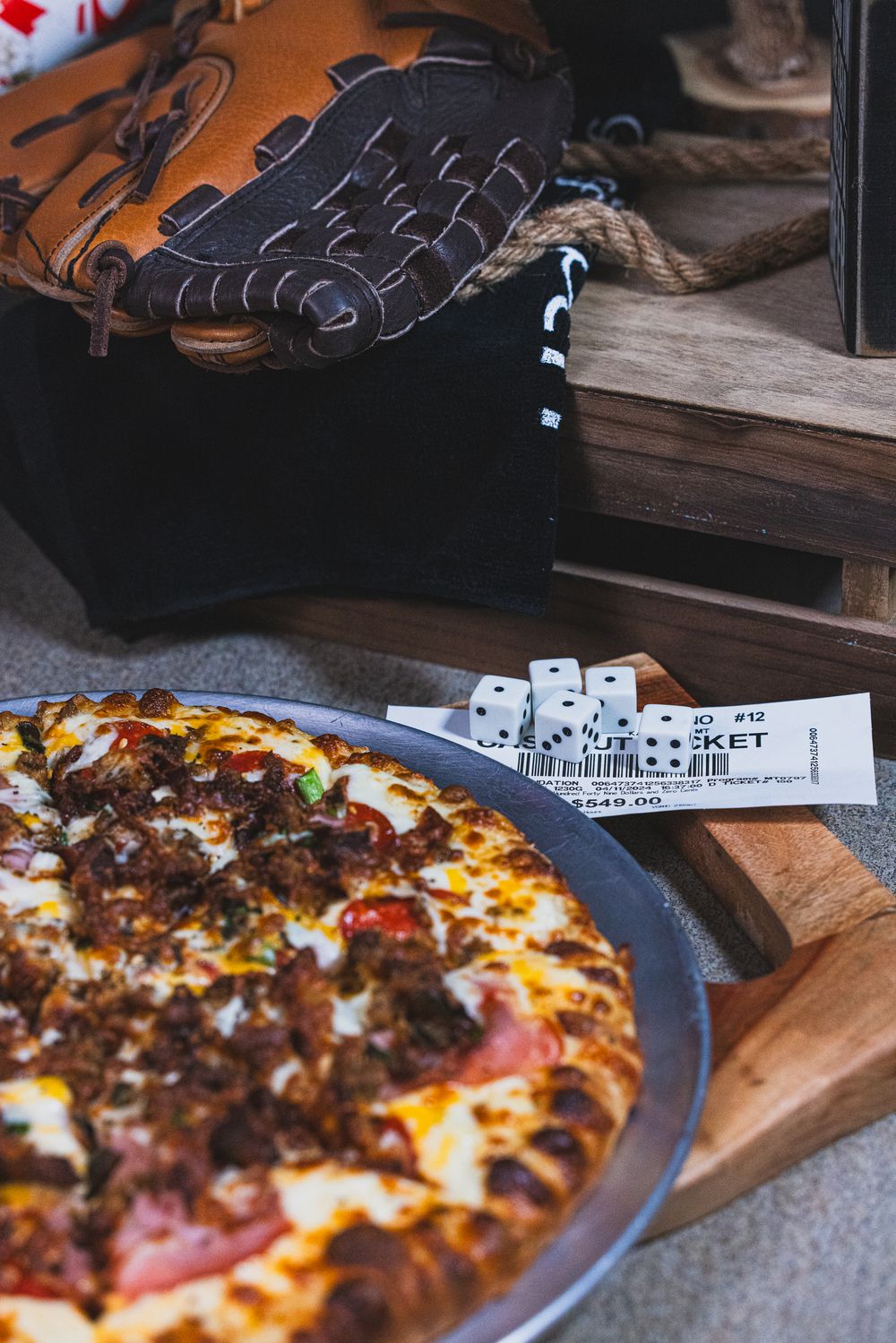 Loaded meat pizza on a wooden board with dice, ticket stubs, and a baseball glove in the background.