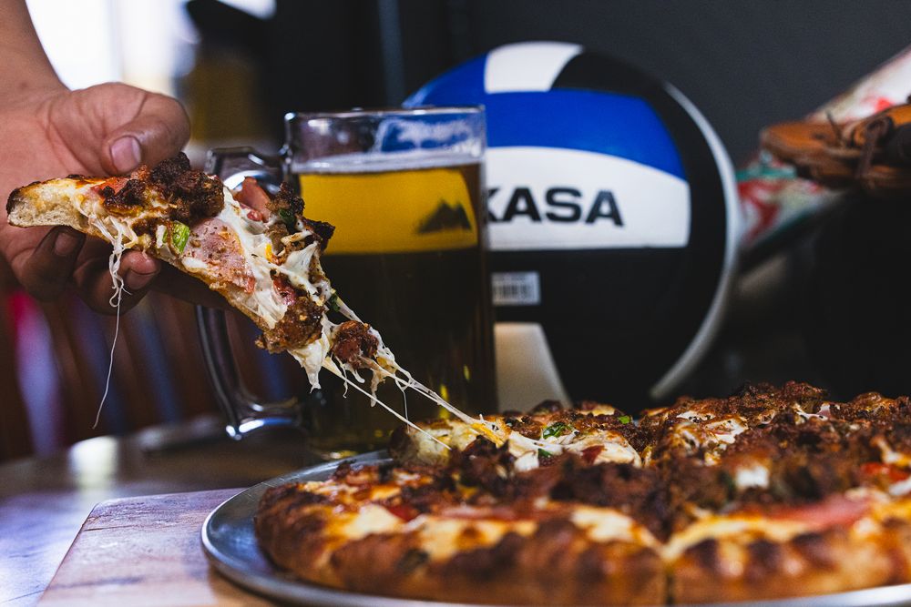 Hand lifting a cheesy pizza slice with a cold beer and volleyball in the background at a sports bar.