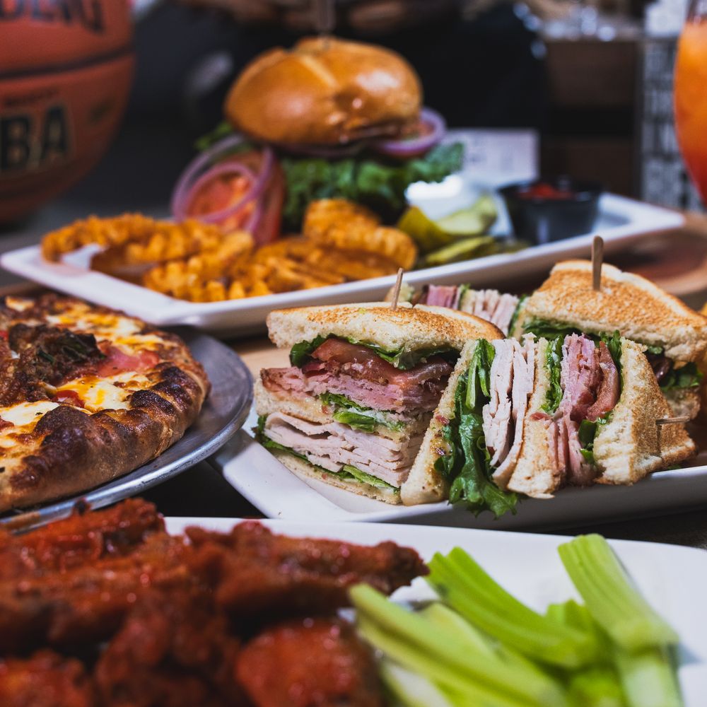 Spread of sports bar food including club sandwich, pizza, wings, burger, and waffle fries on a table.