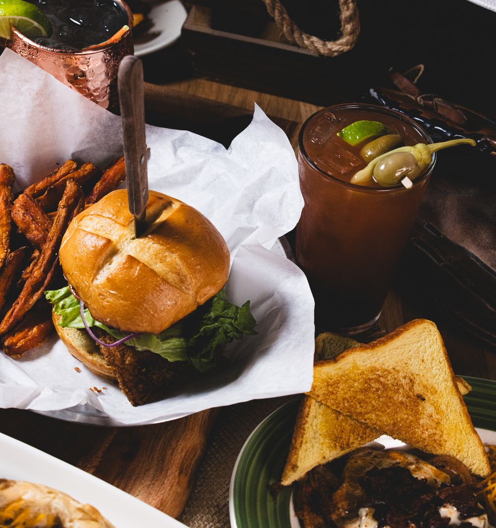 Juicy burger with sweet potato fries, grilled toast, and a spicy Bloody Mary cocktail on a wooden table.