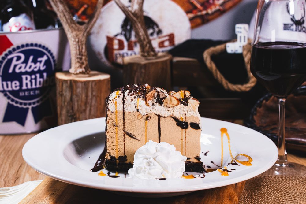 Slice of peanut butter cheesecake with chocolate drizzle and whipped cream on a white plate.