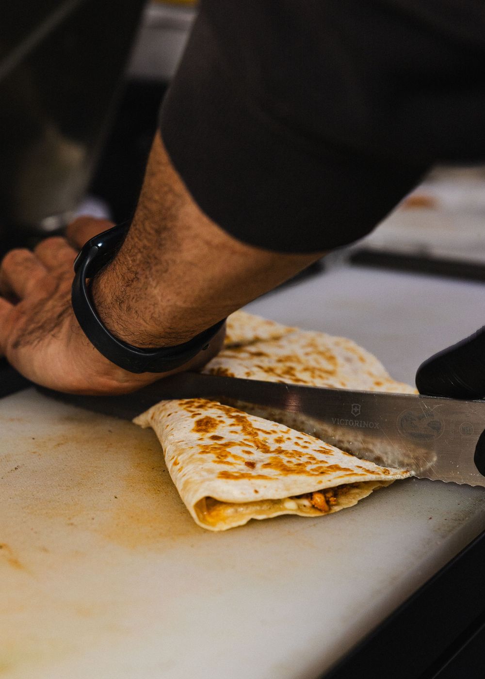 Chef cutting a golden-brown folded quesadilla on a white cutting board with a serrated knife.