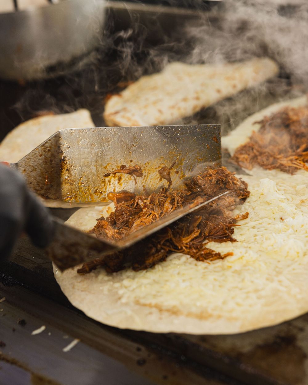 Cook adding seasoned pulled meat filling onto a large flour tortilla on a steaming flat-top grill.