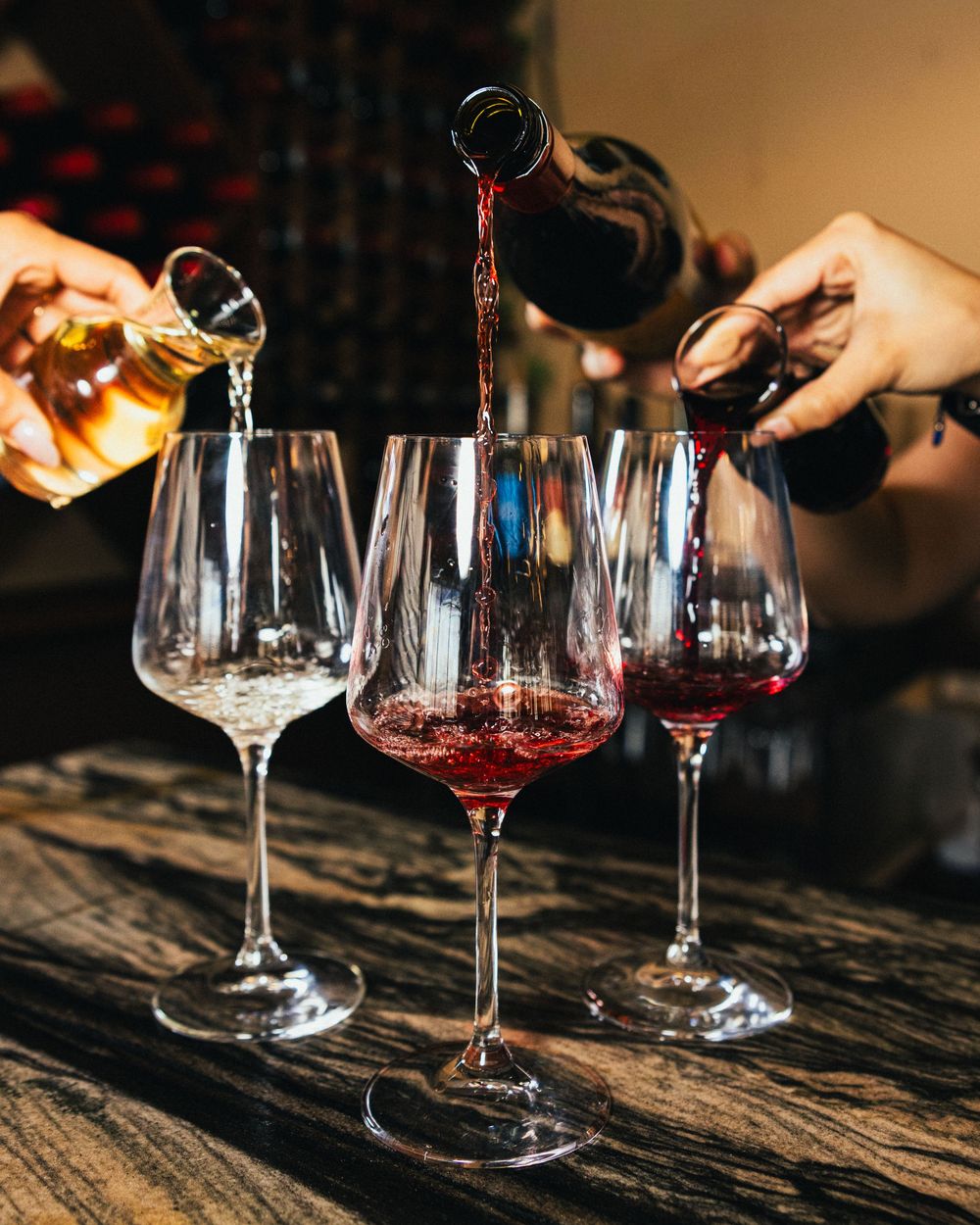 Two people pouring red and white wine into elegant glasses on a rustic wooden table.