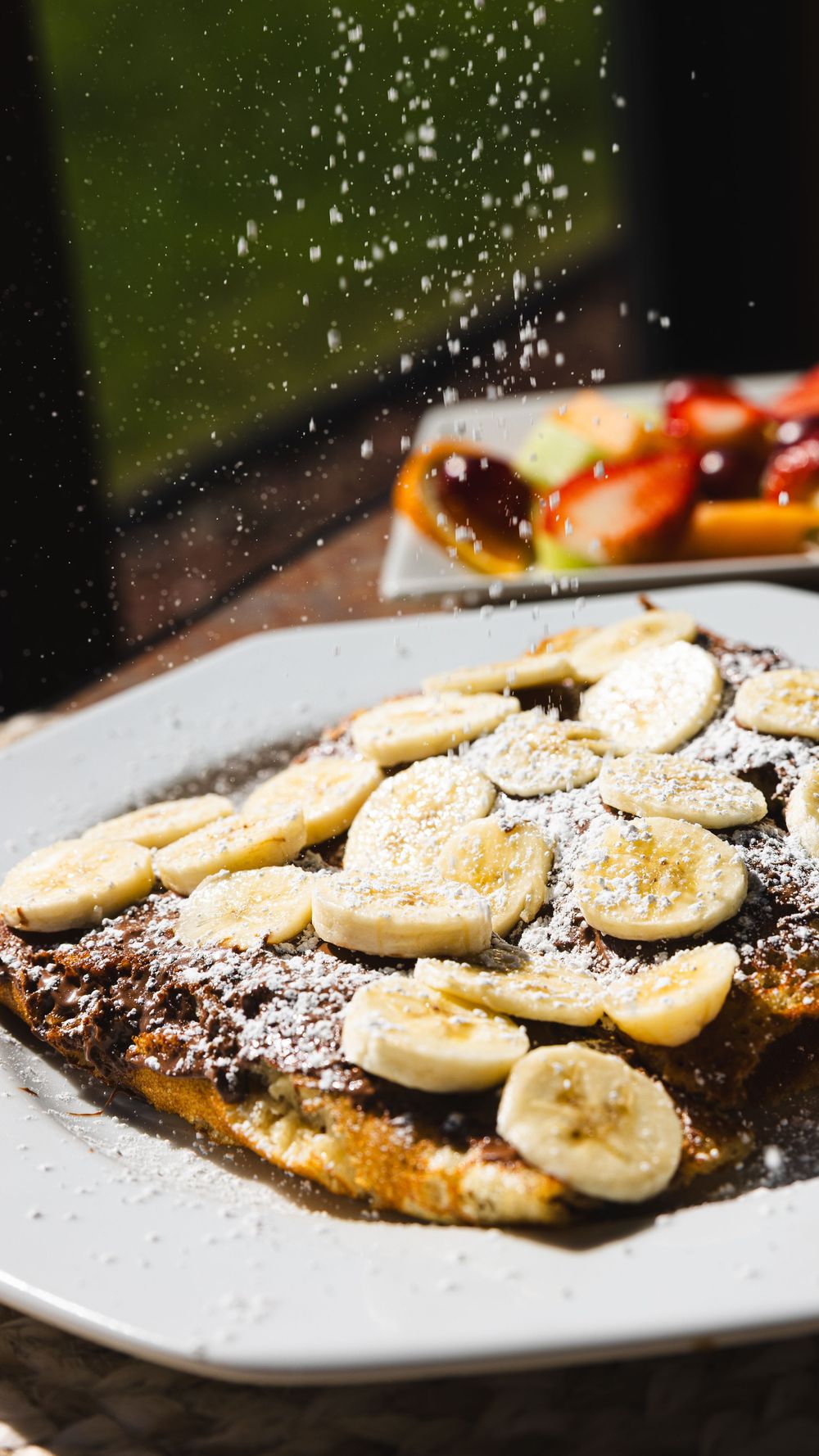 French toast topped with sliced bananas and powdered sugar being sprinkled on white plate.