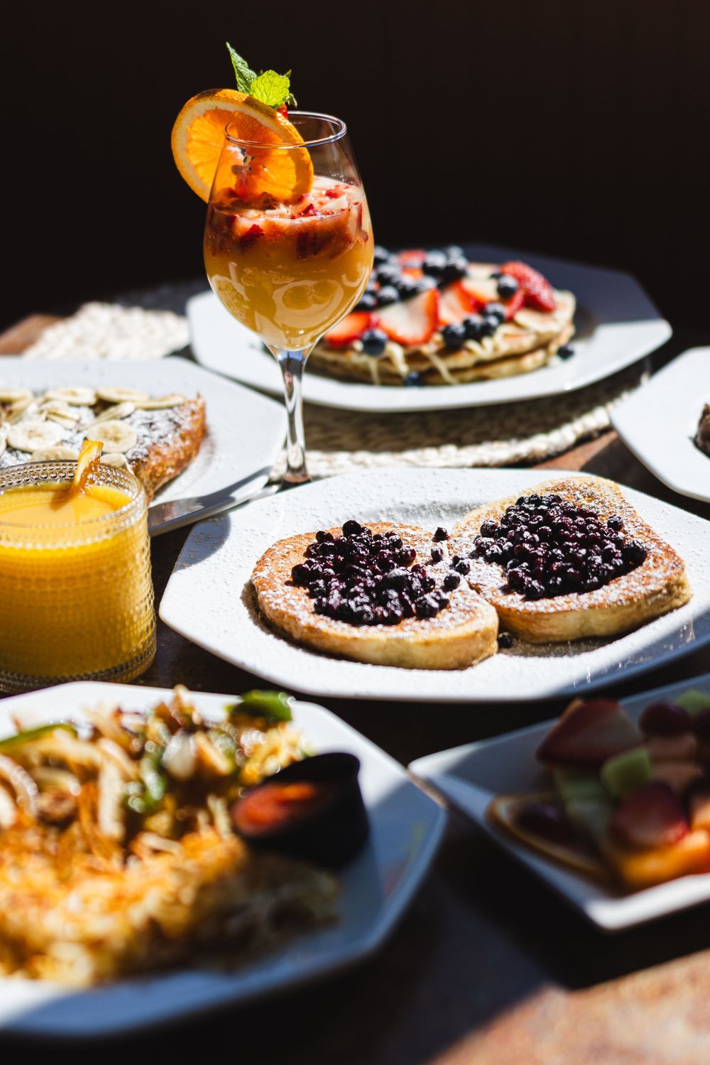 Colorful brunch spread with blueberry toast, fruit pancakes, hash browns, orange juice, and a fresh fruit cocktail drink.