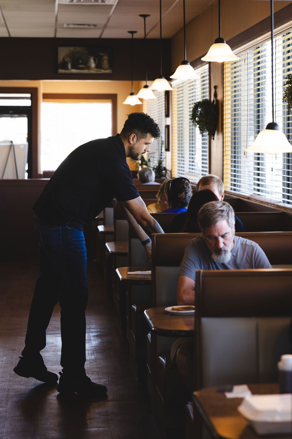Waiter serving customers seated in cozy restaurant booths with warm pendant lighting and wood decor.