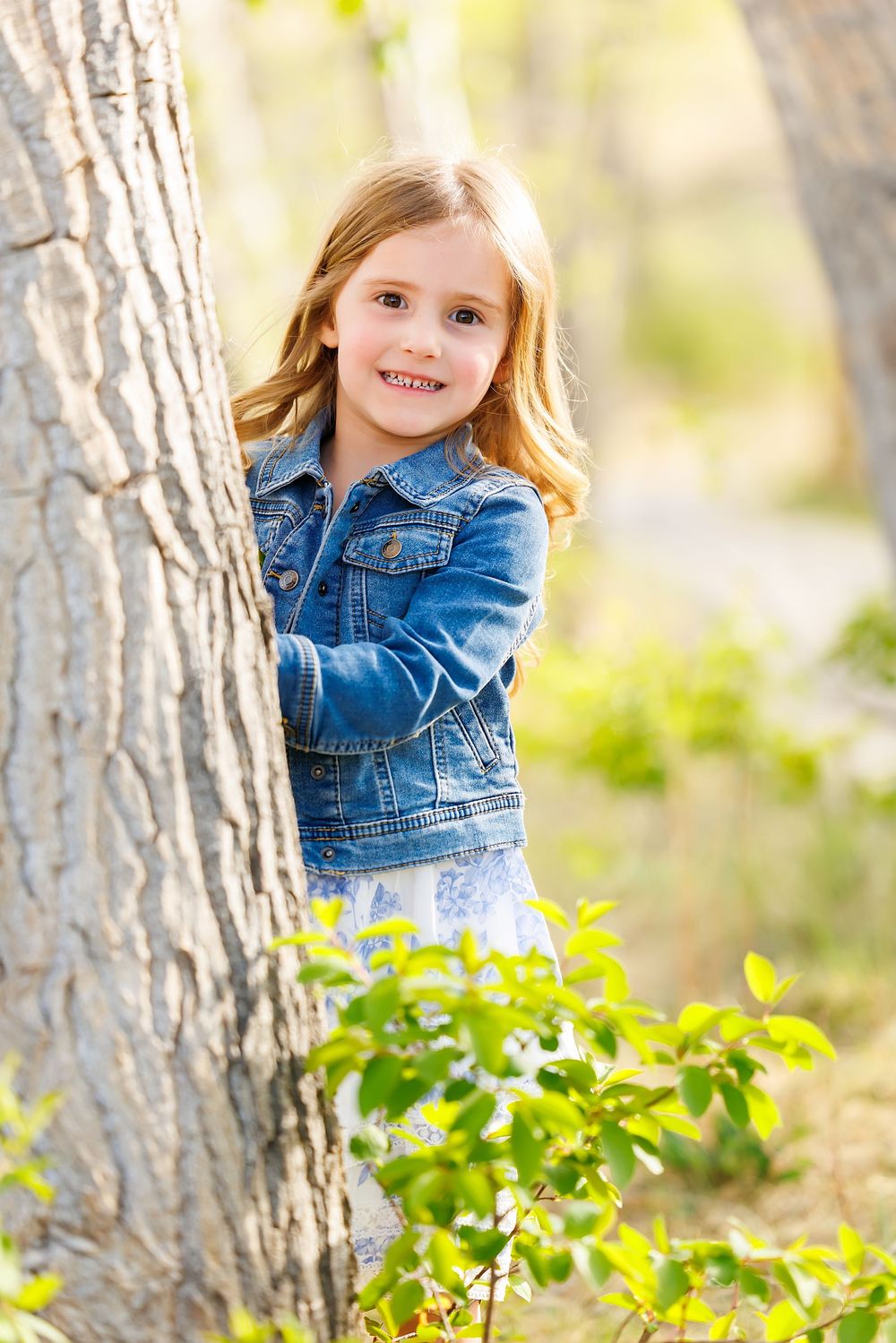 Young girl in denim jacket smiling behind a tree in a sunny outdoor park setting.