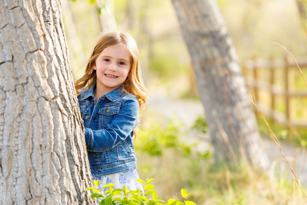 A smiling young girl in a denim jacket peeking out from behind a tree in a sunny outdoor park setting.