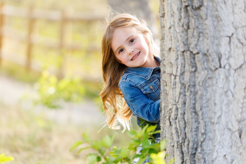 A smiling young girl with long hair peeks from behind a tree trunk wearing a denim jacket in a sunny outdoor setting.