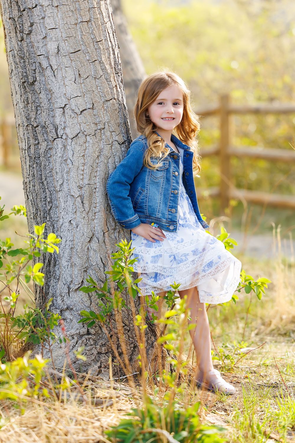 Young girl in denim jacket and white dress leans against tree trunk in sunny outdoor setting.
