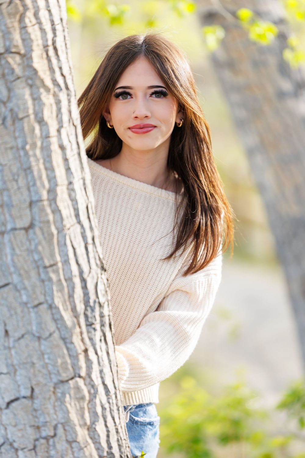 Young brunette woman in cream sweater peeking from behind a tree in a sunny park.