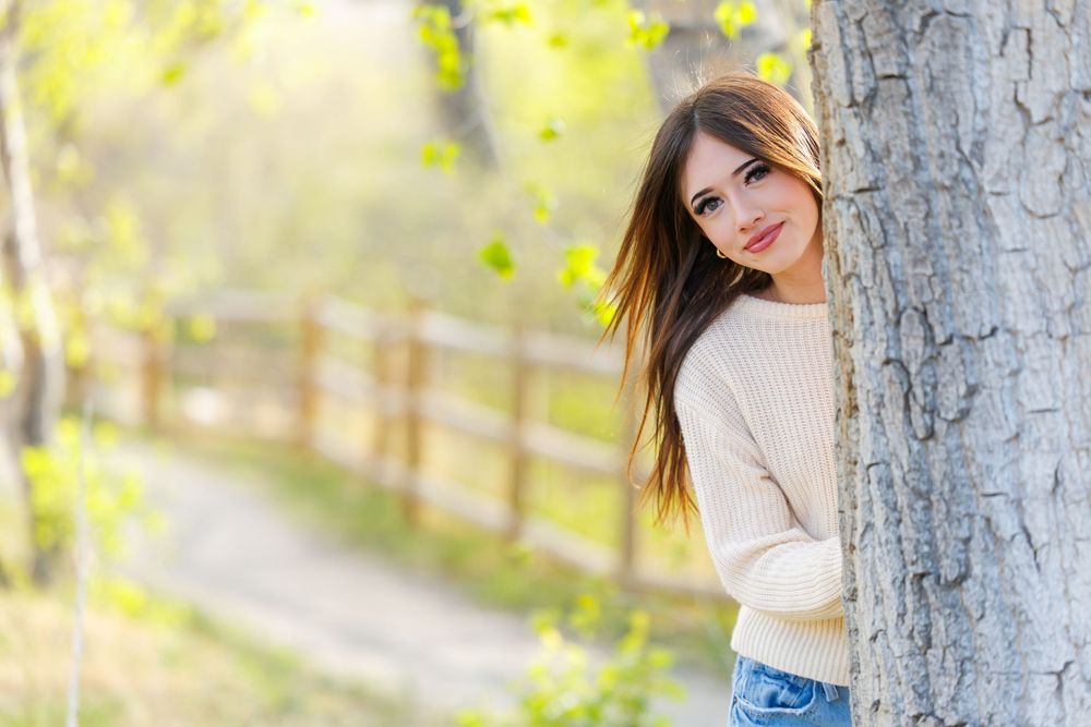 Young woman in cream sweater peeking around a tree trunk in a sunny park setting.