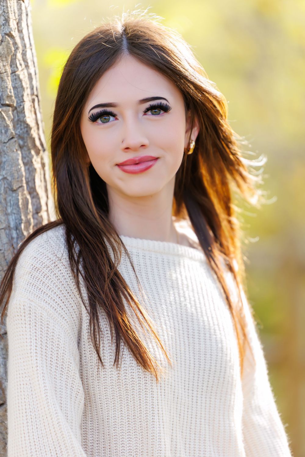 Young woman with long brown hair smiling outdoors in a white knit sweater near a tree in autumn.
