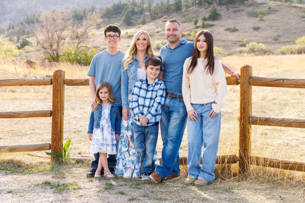Family of six smiling together by a rustic wooden fence in a scenic mountain valley.