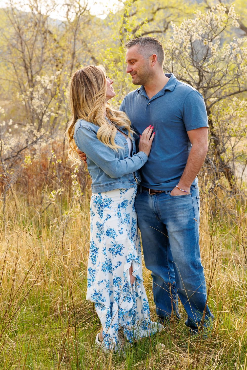 Couple embracing in a golden meadow, woman in floral maxi dress and man in blue polo, surrounded by blooming spring trees.