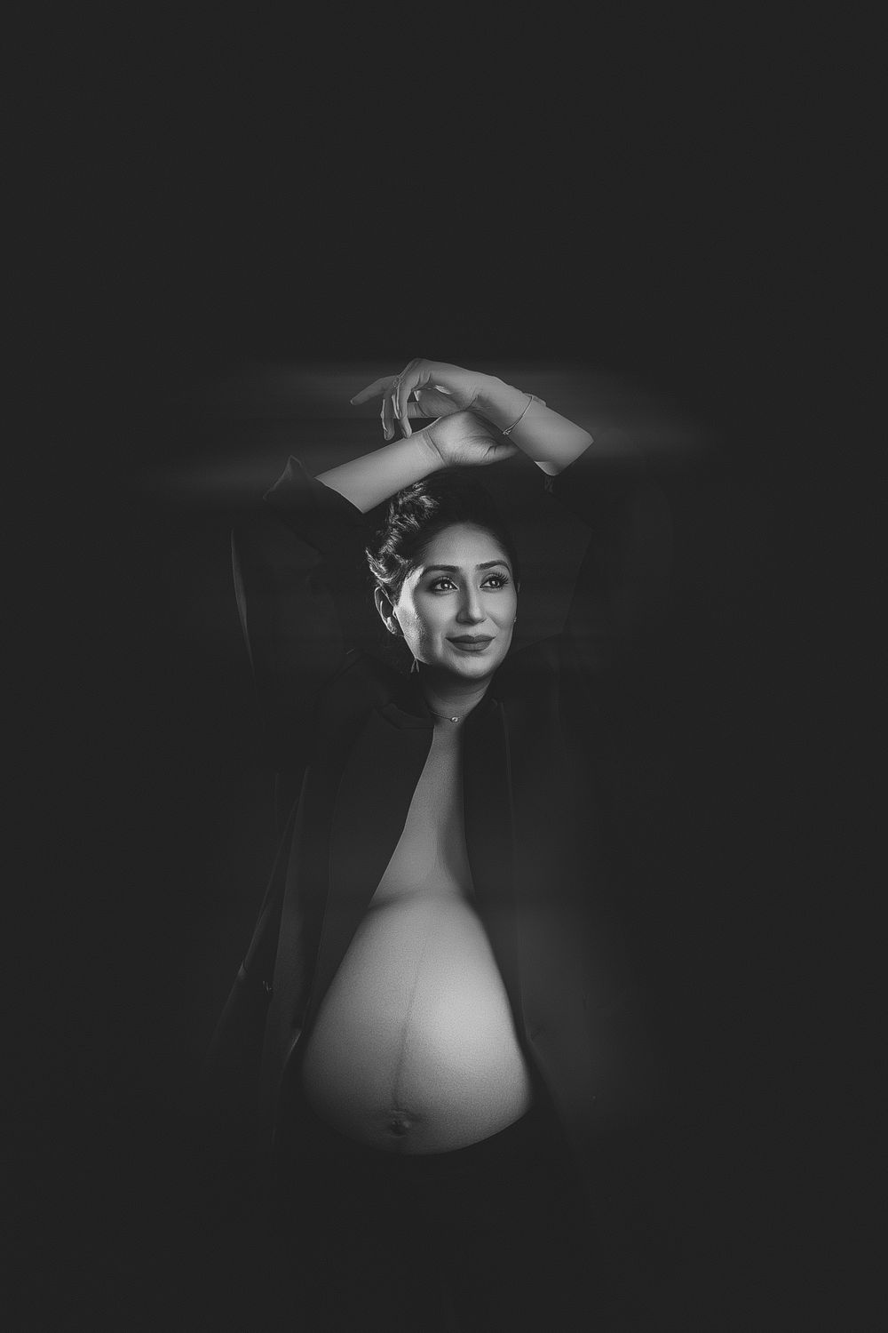 Black and white maternity photo of a pregnant woman posing with arms raised, showcasing her baby bump in dramatic lighting.