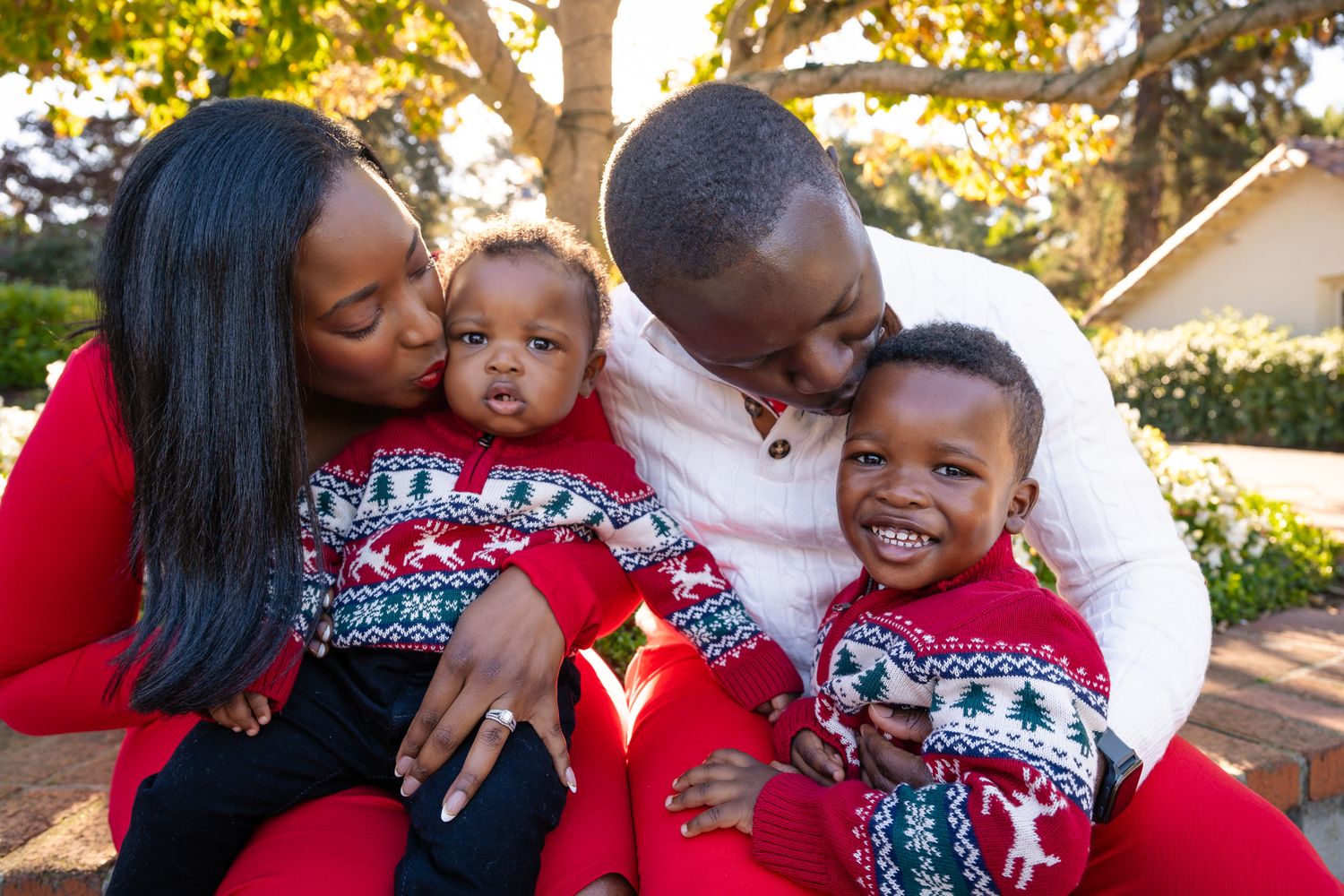 Happy family in matching Christmas sweaters, parents kissing twin babies outdoors during holiday season.