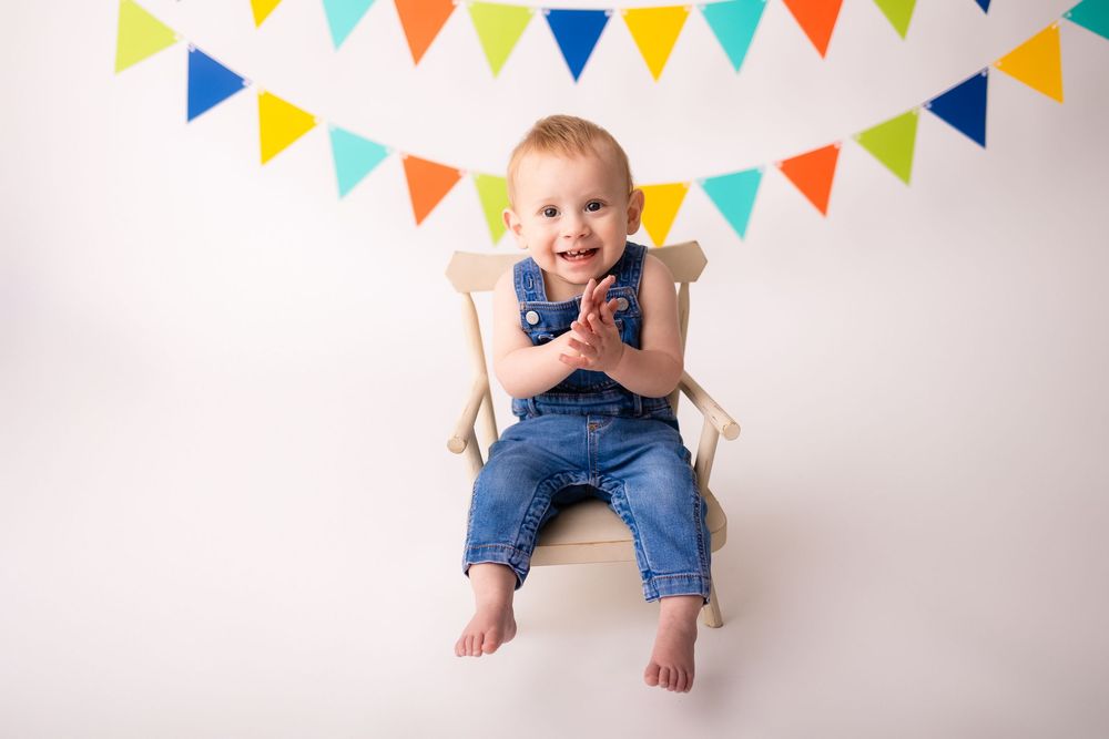 Happy toddler in denim overalls sitting in a wooden chair with colorful birthday bunting banner backdrop.