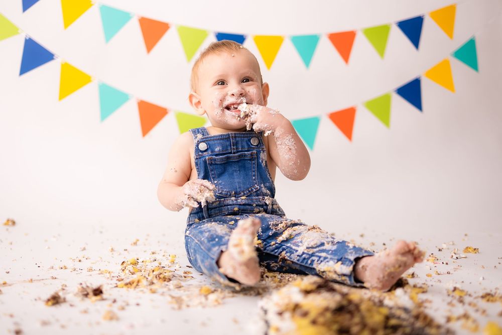 Baby in denim overalls smashing first birthday cake, sitting on white floor with colorful bunting banner backdrop.