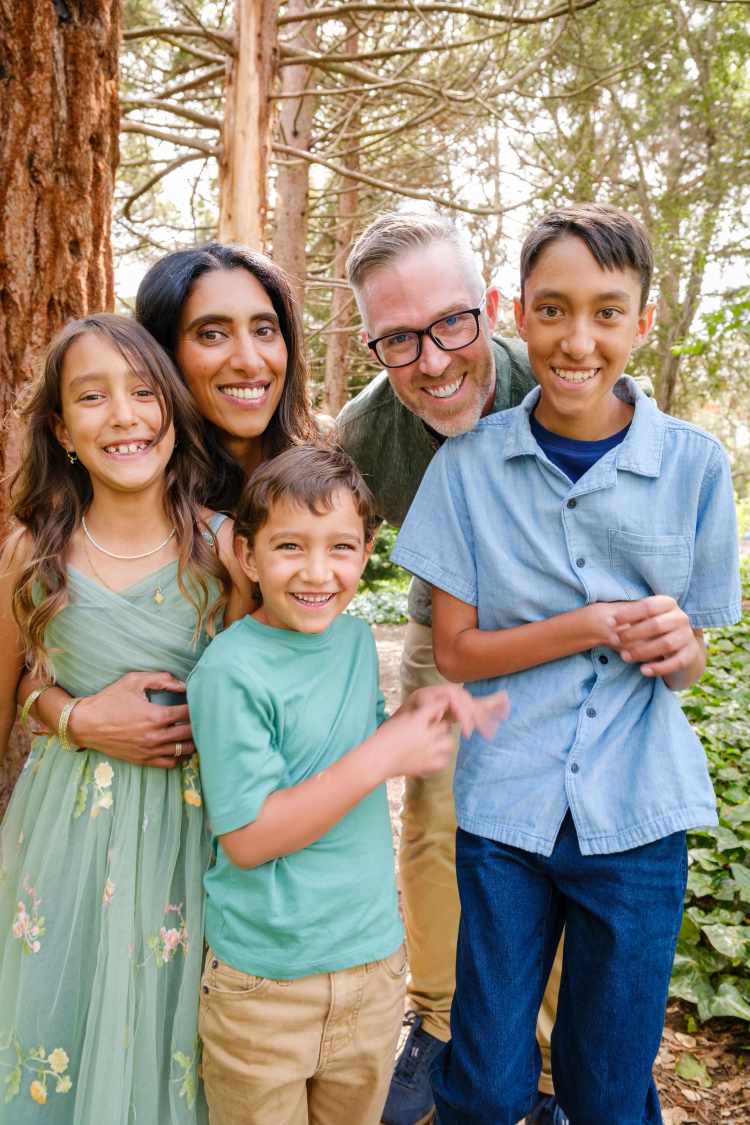 Family of five posing on large twisted oak tree branches in a lush green park setting.