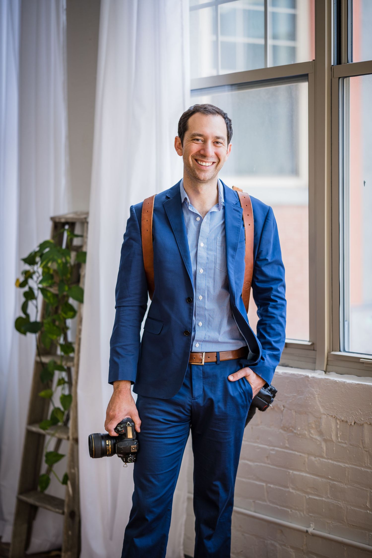 Smiling man in blue suit holding DSLR camera with leather strap, standing by large window in bright studio space.