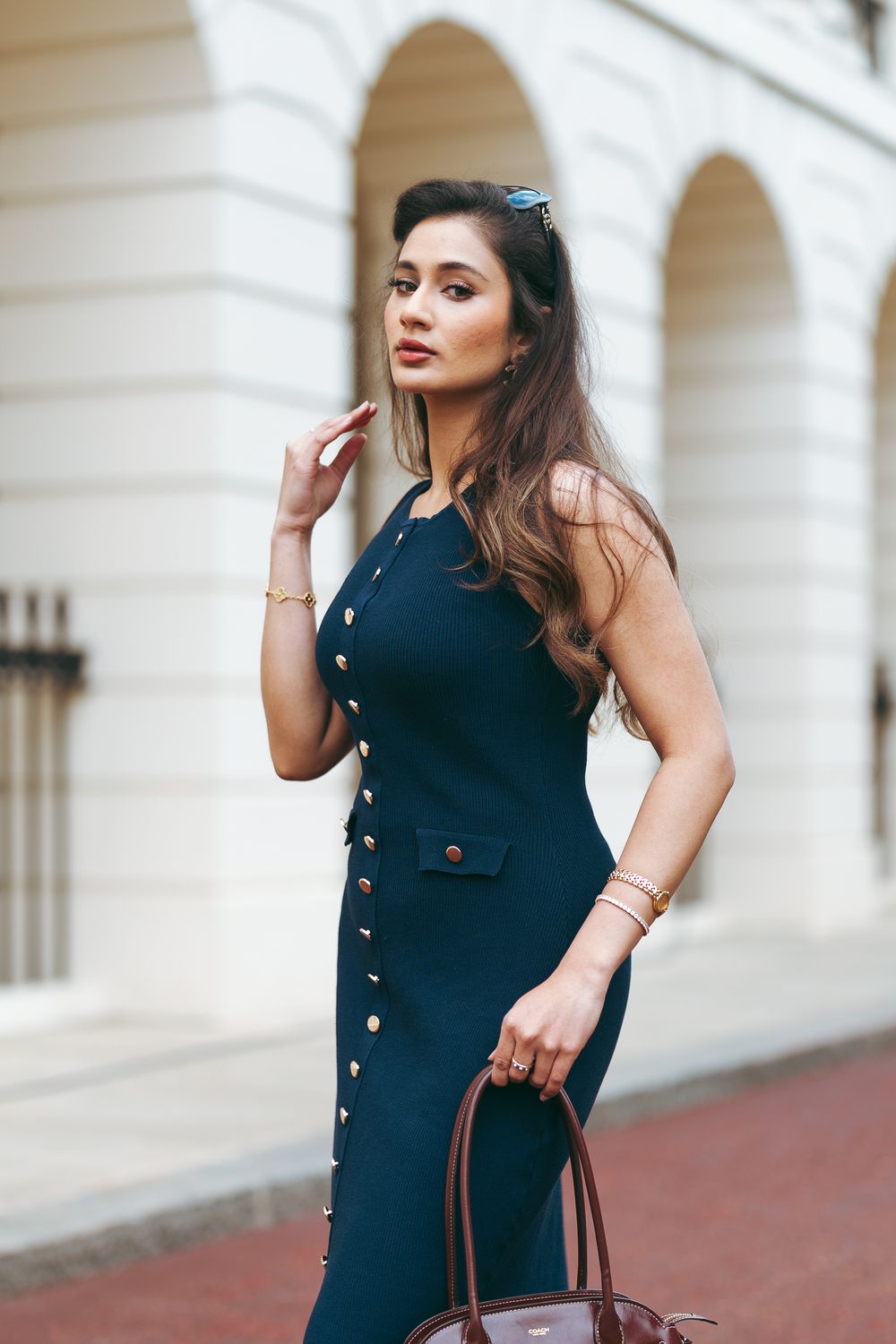 Elegant woman in navy blue button-front dress holding brown handbag, posing outdoors near white arched building.
