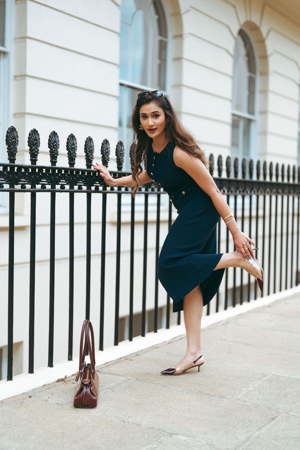 Woman in dark teal dress posing by black iron fence on city street, holding shoe with brown handbag nearby.