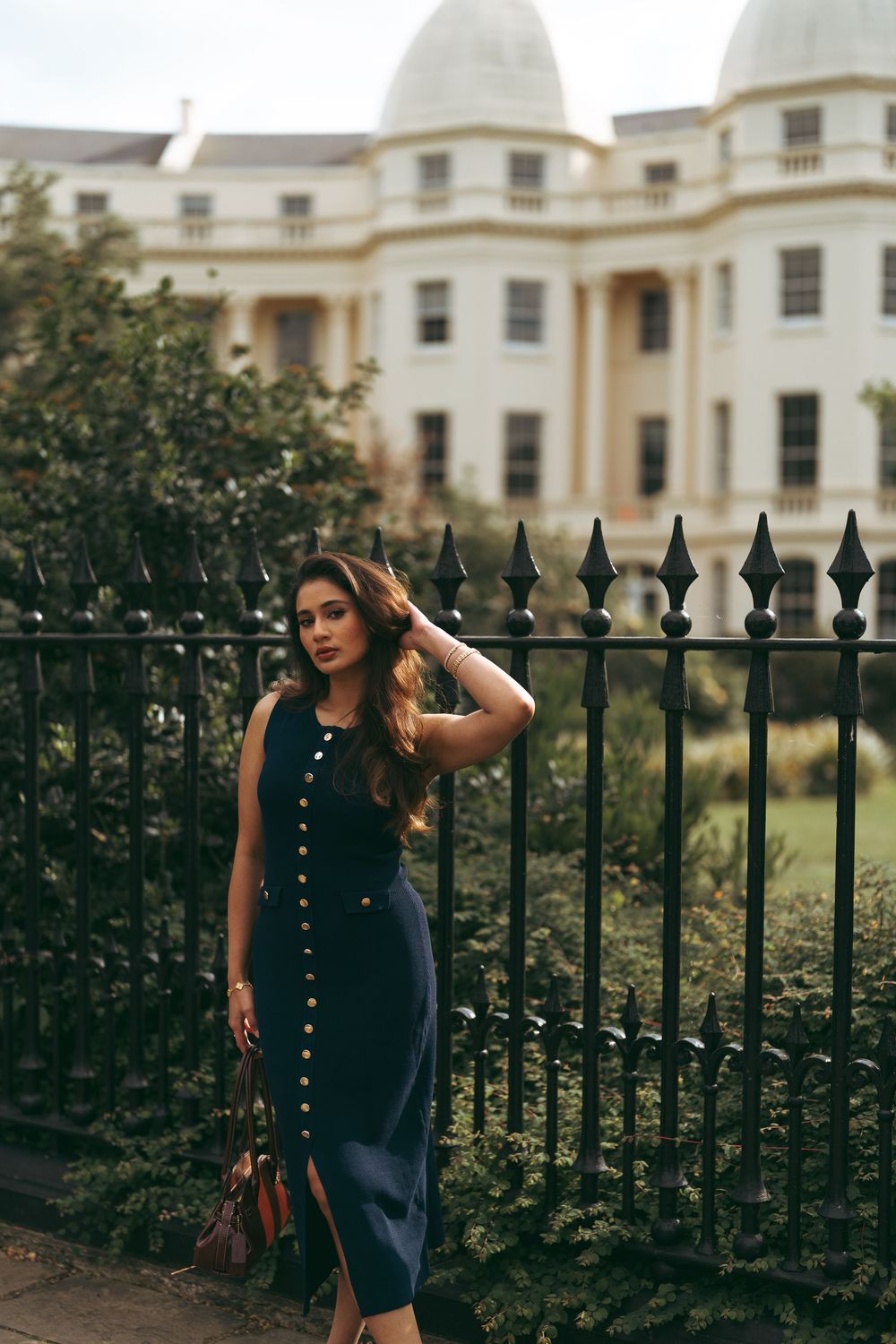 Woman in navy midi dress with gold buttons poses by iron fence with grand white building backdrop.