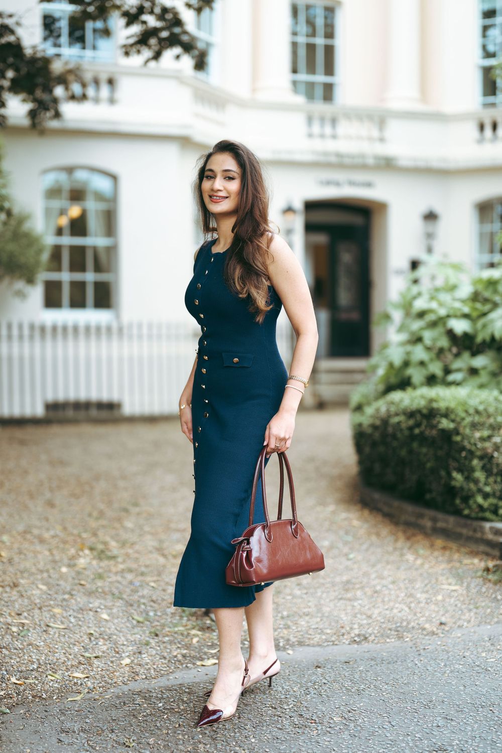 Elegant woman in navy sleeveless midi dress carries burgundy handbag outside white Georgian building.
