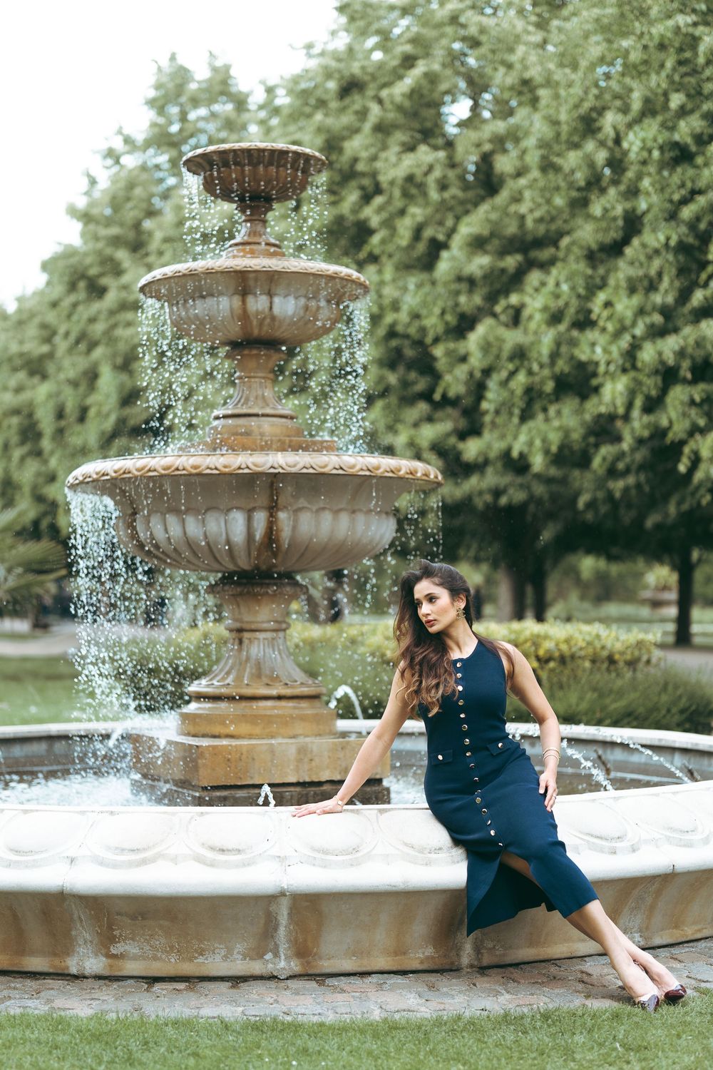 Woman in navy dress posing by a tiered stone fountain in a lush green park.