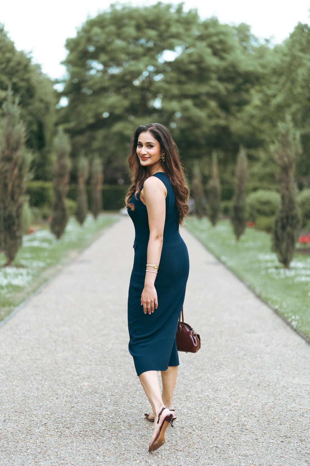 Woman in navy midi dress walking through formal garden path lined with trees, carrying brown handbag.