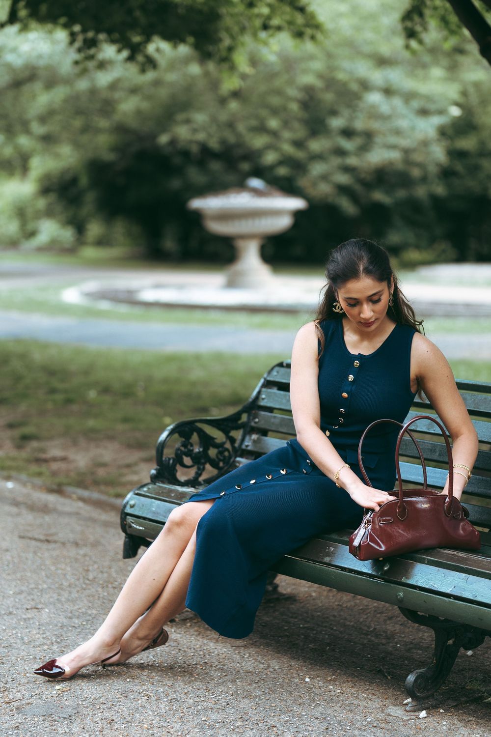 Elegant woman in navy button-front dress sitting on park bench with brown leather handbag.