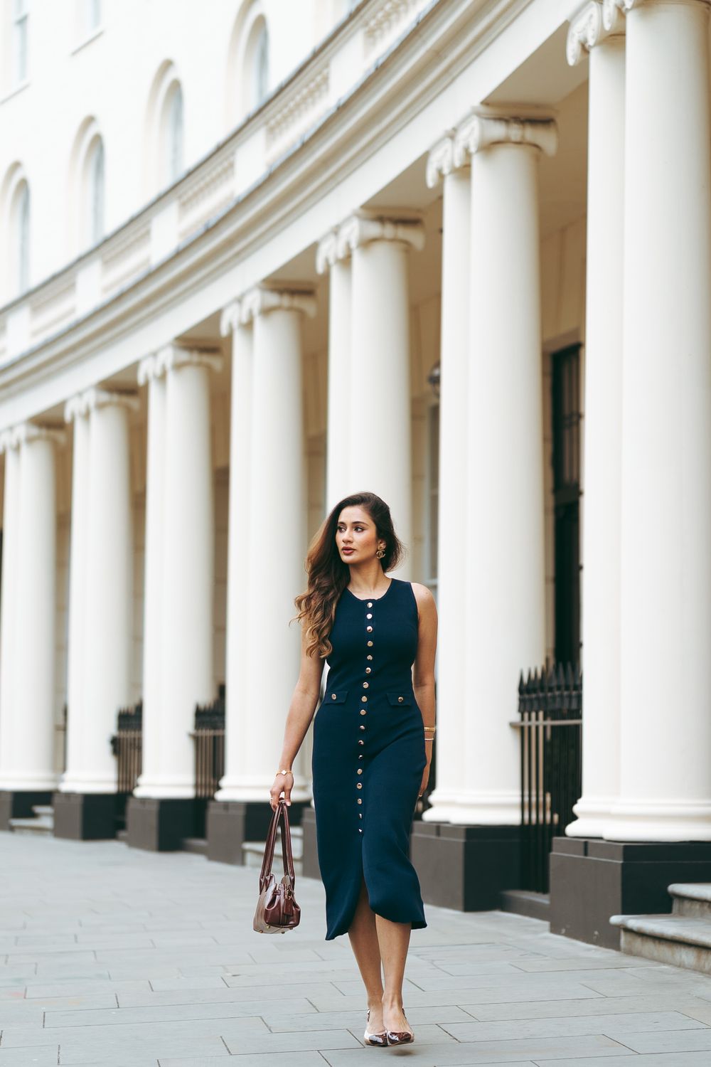 Woman in navy sleeveless midi dress walking past white neoclassical columns carrying brown bag.