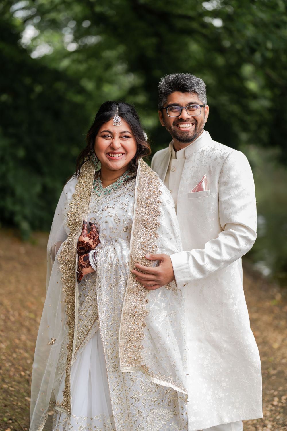 Indian couple in white traditional wedding attire smiling outdoors, bride in embroidered lehenga with dupatta, groom in sherwani.
